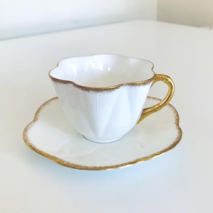May include: A white teacup and saucer set with a scalloped edge and gold trim. The teacup has a gold handle and a fluted design. The saucer is shaped like a flower. A classic tea set.