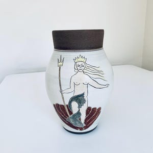 May include: White ceramic vase with a dark brown rim. The vase features a hand-drawn illustration of a mermaid holding a trident, seated on a red and brown seashell. The mermaid wears a crown. The vase is a decorative item.