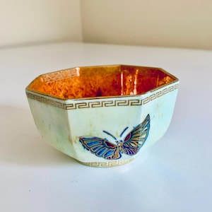 May include: An octagonal bowl with a light green exterior and a vibrant orange interior. The bowl has a gold Greek key pattern around the rim and base, and a colourful butterfly design on the front. A decorative piece.