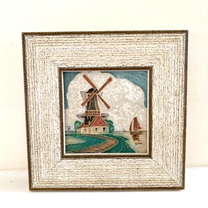 May include: A framed artwork depicting a windmill with a red roof and a blue sky with white clouds. A sailboat is in the water in the foreground.