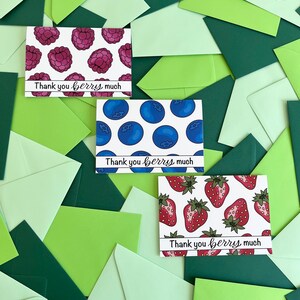 Puede incluir: Tarjetas de felicitación con ilustraciones de bayas, incluyendo frambuesas, arándanos y fresas, están dispersas entre sobres verdes. Cada tarjeta tiene la frase "Thank you berry much" impresa. Las tarjetas y los sobres son en tonos verdes.
