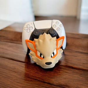 Może przedstawiać: Biały kontroler PlayStation 5 z brązową i pomarańczową głową postaci przymocowaną do przodu. Postać ma spiczasty pysk, duże oczy i małe usta.