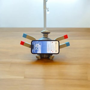 May include: A grey phone stand with a smartphone displaying notifications. The stand has a robot design with arms featuring red and blue accents. The phone screen shows the time as 7:24.