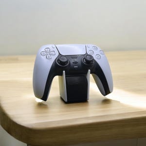 May include: A white and black wireless game controller on a black charging stand. The controller features a symmetrical design with buttons and joysticks. The stand is rectangular and supports the controller.