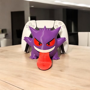 Può includere: Una statuetta di Ghost Pokemon stampata in 3D di colore viola e rosso che tiene un controller PlayStation 5 bianco. La statuetta ha un ampio sorriso e una grande lingua rossa che sporge.