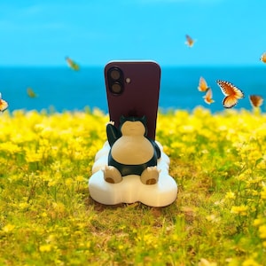 Op de afbeelding: Een bordeauxrode smartphone wordt vastgehouden door een witte, wolk-vormige telefoonstandaard met een cartoonfiguur. De standaard staat in een veld met gele bloemen met vlinders op de achtergrond.