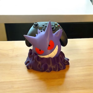 May include: A purple Gengar-themed controller stand with red eyes and a white grin, holding a black game controller. The stand has a textured, dark purple base, and is on a wooden surface.