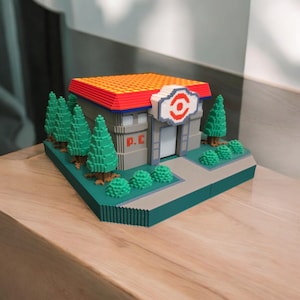 May include: A 3D printed model of a Pokemon Center building with a red roof, a white sign with a red eye, and the text "P.C." on the side. The building is surrounded by green trees and bushes.