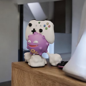 May include: A white Xbox controller is paired with a purple cartoon character figurine. The figurine has a smiling face and a skull and crossbones design. The controller has coloured buttons and a black directional pad.
