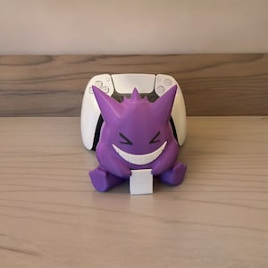 Könnte beinhalten: Eine lila Gengar-Figur mit breitem Grinsen, die einen kleinen weißen Block hält, vor einem weißen Gamecontroller. Die Figur hat spitze Stacheln auf dem Kopf und sitzt auf einer hellen Holzoberfläche.