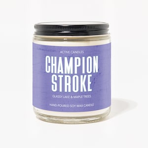 May include: A glass jar candle with a purple label that reads "ACTIVE CANDLES CHAMPION STROKE GLASSY LAKE & MAPLE TREES HAND-POURED SOY WAX CANDLE".