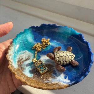 May include: A round resin trinket dish featuring a blue, turquoise, and sandy brown ocean-inspired design with a gold-tone chain necklace with a rectangular pendant labelled The Empress and a small brown and tan sea turtle figurine on top.