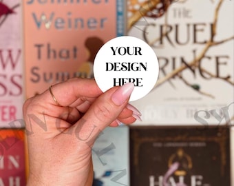 May include: A white round sticker with black text that reads "YOUR DESIGN HERE" is being held by a hand in front of a bookshelf with various book covers.