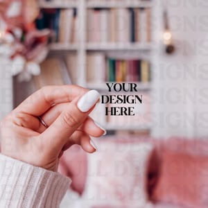 May include: A hand holding a blank white card with the text "YOUR DESIGN HERE" in black. The hand is wearing a white sweater and has a ring on the middle finger. The background is blurred and shows a bookshelf and a pink couch.