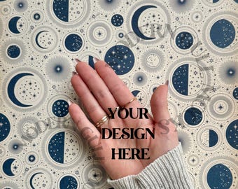 May include: A celestial-themed design featuring navy blue crescent moons, stars, and phases of the moon on a cream background. The words "YOUR DESIGN HERE" are printed on the palm of a hand, with gold rings on the fingers. The person is wearing a gray sweater.