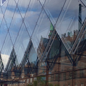 Puede incluir: Un edificio con una fachada de vidrio que refleja el cielo y otros edificios. Los paneles de vidrio están dispuestos en forma de diamante, creando un diseño geométrico.