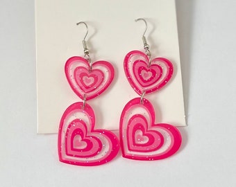 Peut inclure: Une paire de boucles d'oreilles pendantes en forme de cœur rose avec des tourbillons blancs et des paillettes. Les boucles d'oreilles ont des crochets argentés.