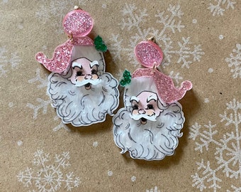 May include: A pair of pink glitter Santa Claus earrings. The earrings are made of clear acrylic with a white and black illustration of Santa's face. The Santa hats are pink glitter.
