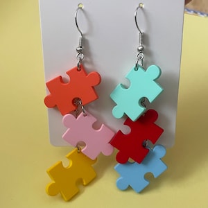 May include: A pair of dangle earrings featuring five colorful puzzle piece charms. The charms are orange, pink, yellow, blue, and red.