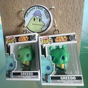 May include: A pair of handmade earrings featuring miniature Greedo figures from Star Wars. The earrings are attached to a card with a "Grumpy Frog Press" logo. The Greedo figures are green with yellow accents.