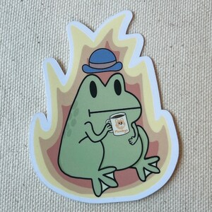 May include: A green frog wearing a blue hat and holding a coffee mug with the word "Mondays" on it. The frog is sitting on a yellow and orange background.