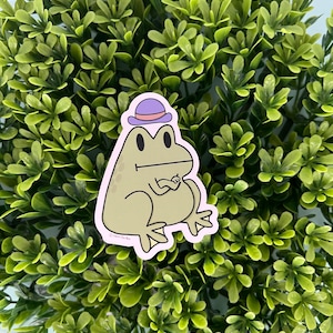 May include: A green frog wearing a purple hat with a white band sits on a green leafy background. The frog has a white outline and is looking to the right.
