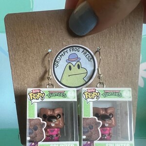 May include: A pair of handmade earrings featuring miniature Splinter figures from the Teenage Mutant Ninja Turtles. Each figure is displayed in a clear plastic box, with the words "Bitty Pop! Turtles" and "Splinter" visible. The earrings are attached to a brown card.