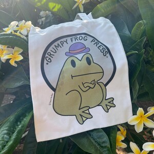 May include: White tote bag featuring a green frog wearing a purple and orange hat. The bag is printed with the text "Grumpy Frog Press."