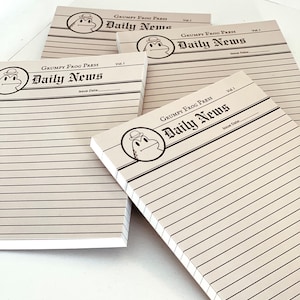 May include: Four brown and white lined notepad paper pads with a cartoon frog logo and the text "Grumpy Frog Press Daily News" and "Vol. 1" printed on the front.