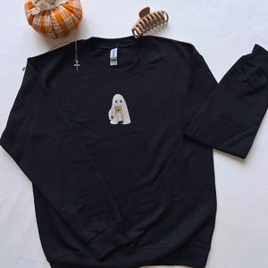 May include: Black sweatshirt featuring an embroidered ghost holding a coffee cup. The ghost is white with a simple design. The sweatshirt has long sleeves and a crew neck. A small pumpkin and hair clip are in the background.