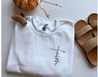 Puede incluir: Camiseta blanca con la palabra "faith" escrita verticalmente en letra negra, con una cruz en la parte superior de la palabra.