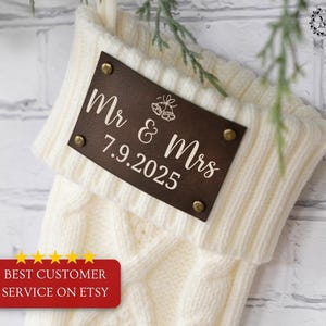 May include: Cream-colored cable knit Christmas stocking with a brown leather tag. The tag is inscribed with "Mr & Mrs" and the date "7.9.2025". The tag is secured with brass rivets. A red banner at the bottom reads "BEST CUSTOMER SERVICE ON ETSY".