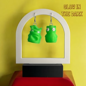 May include: A pair of green glow-in-the-dark earrings featuring cartoon characters from the animated series "Aqua Teen Hunger Force". The earrings are hanging from a white arch-shaped display stand. "GLOW IN THE DARK" is written in orange text on a yellow background.