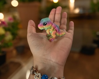 May include: A hand holding a small, rainbow-colored dinosaur toy. The hand is wearing a silver charm bracelet with blue and gold beads.