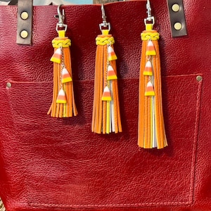 May include: A red leather tote bag with three decorative keychains. Each keychain features a long orange tassel with yellow and white accents, and candy corn charms. The bag has a front pocket and brown leather handles.