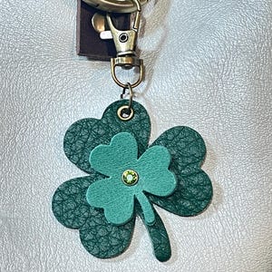 May include: A green leather four-leaf clover keychain with a sparkling green rhinestone in the center. The keychain is attached to a gold-toned metal ring and a lobster clasp.