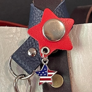 May include: A red star-shaped leather keychain charm with a silver button and a small star charm featuring the American flag. The charm is attached to a silver clasp and leather straps in navy blue and brown.