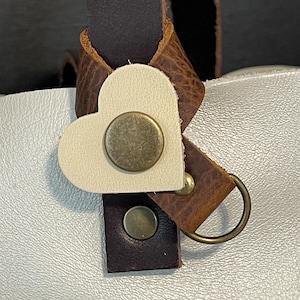 May include: A white leather heart-shaped charm with a brass button and ring attached to a brown leather strap.
