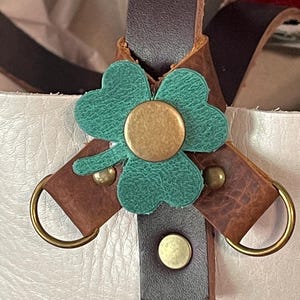 May include: Close-up of a leather bag detail featuring a teal shamrock with a gold center, attached to brown leather straps with gold metal hardware. The bag has a white leather exterior.