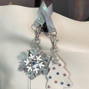 May include: A white leather tote bag with a silver iridescent strap and two charms. One charm is a snowflake with blue rhinestones and the other is a Christmas tree with blue and pink rhinestones.