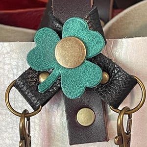 May include: Close-up of a leather bag strap with a teal shamrock accent. The shamrock is secured with a brass-colored rivet in the center. The strap features black and brown leather with brass-colored hardware.