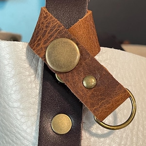 May include: Close-up of a leather bag strap with a brass-colored metal ring and rivets. The strap is brown and tan with a textured appearance. The bag appears to be white leather.