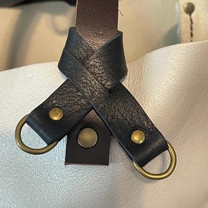 May include: Close-up of a leather tote bag handle with a brown leather strap and black leather straps with brass accents.