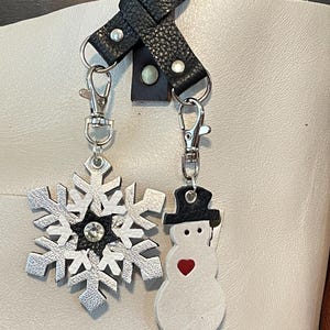 May include: A white leather tote bag with a black leather strap and two charms. One charm is a silver snowflake with a rhinestone in the center. The other charm is a white snowman with a black hat and a red heart.