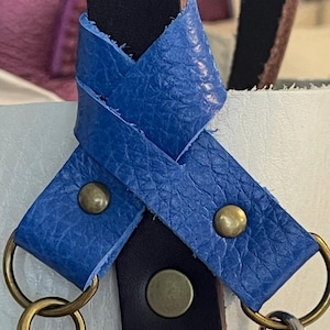 May include: Close-up of a leather bag strap with blue and black leather accents. The blue leather is secured with brass rivets and attached to metal rings and clasps. The strap is designed for a bag or purse.