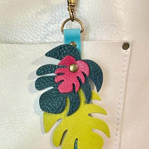 May include: A tropical-themed keychain featuring a layered leather design. The keychain showcases a vibrant yellow, teal, and dark green monstera leaf with a pink flower accent. It includes a gold-colored clasp and a teal leather strap.