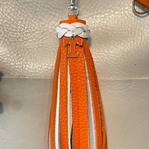 May include: Orange and white tassel keychain with a silver clasp. The tassel features alternating orange, white, and gray leather strips. A silver charm with an orange "T" is attached to the top, and a white braided leather detail is above the charm.