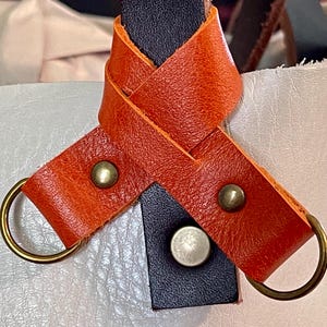 May include: Close-up of a leather bag strap detail. The strap features orange leather with brass rivets and gold-colored metal rings. A black leather piece is also visible, with a silver rivet. The bag appears to be white or cream-colored.
