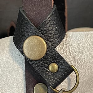 May include: Close-up of a black leather strap with a brass buckle and a silver key ring. The strap is attached to a white leather bag.