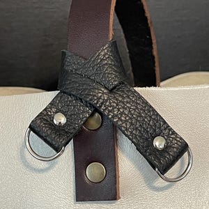 May include: Close-up of a white leather tote bag with a dark brown handle and black leather accents. The handle is a dark brown leather strap. The black leather accents are attached with silver-colored rivets and metal rings.